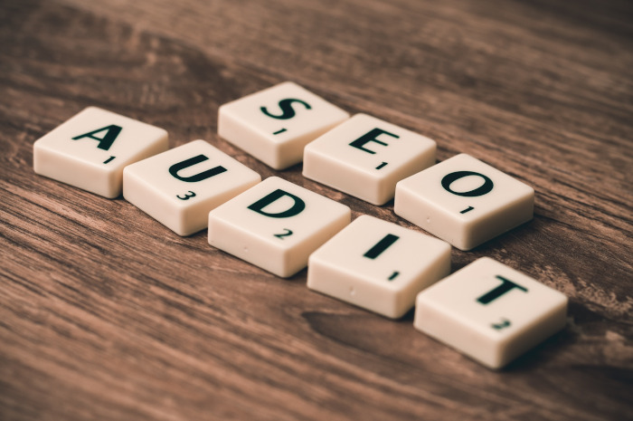 Scrabble-Steine mit schwarzen Buchstaben SEO AUDIT liegen auf einem braunen Holztisch.