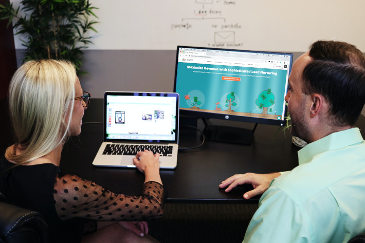 Mann und Frau am Tisch sitzend betrachten Webdesign, vor Ihnen ein Desktop-Bildschirm und Laptop